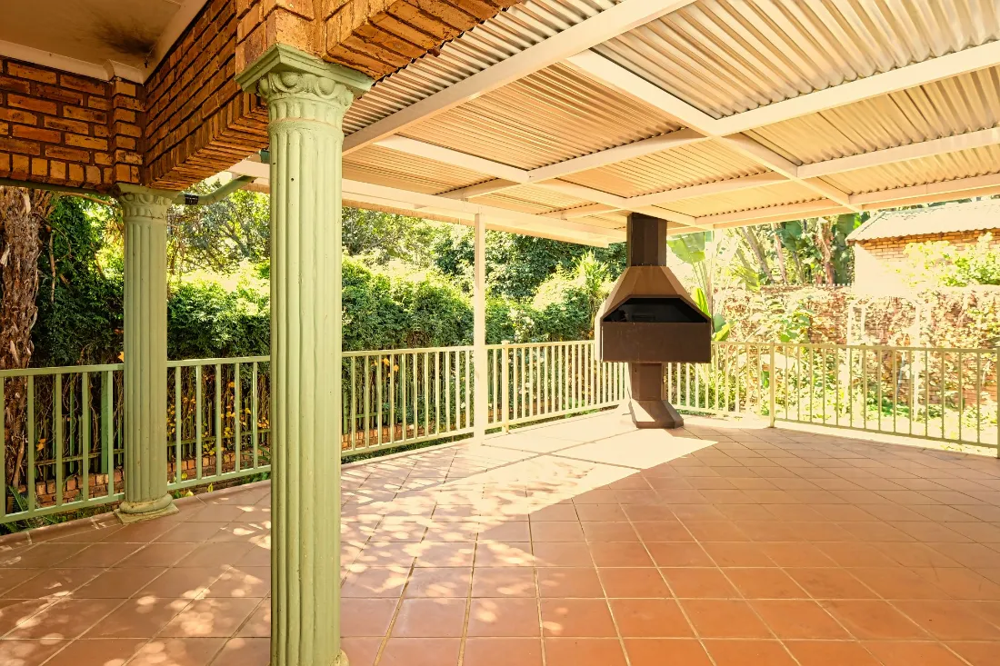 Porche cubierto con pérgola, suelo de barro y barbacoa de obra, con el jardín detrás
