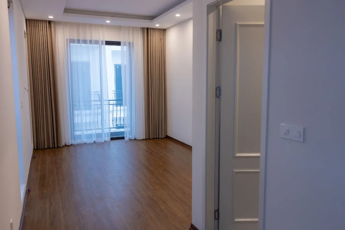 Salón vacío con suelo de madera y puerta balconera con cortinas